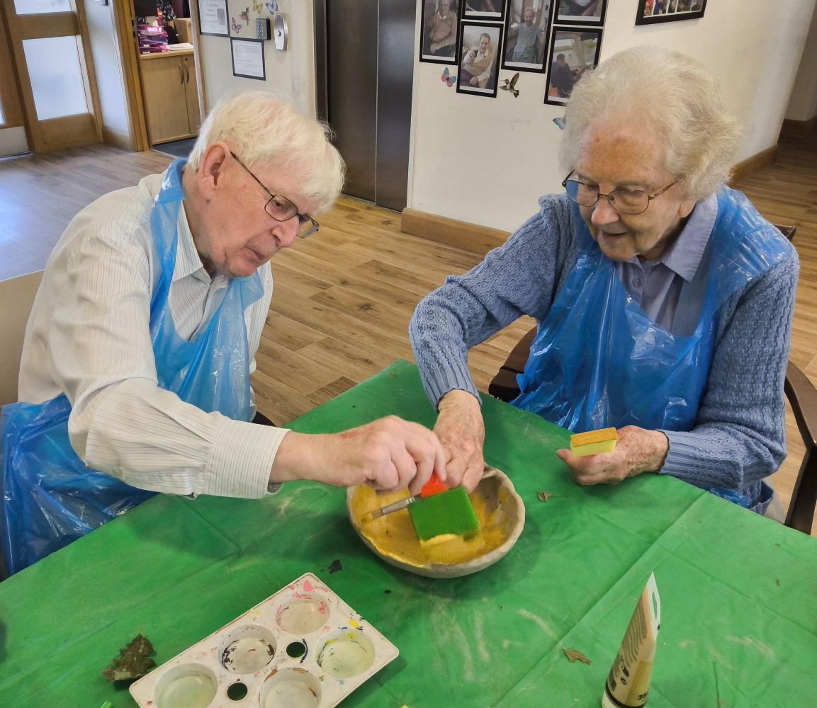 A couple seated side by side, wearing blue plastic aprons, applying gold acrylic paint to the inside of a clay bowl