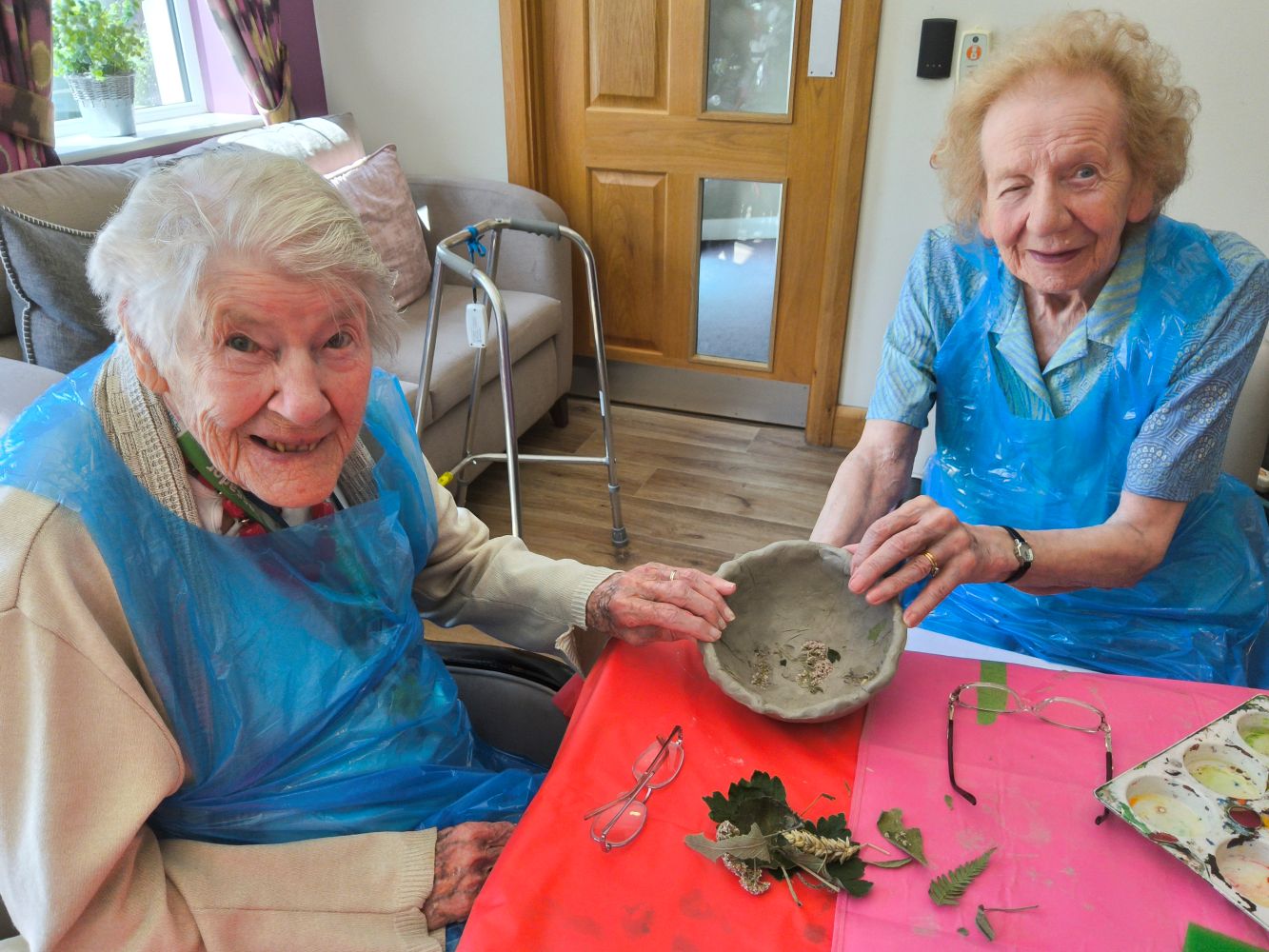 Two women position their clay bowl to demonstrate that they have picked out most of the leaves - there's a small pile in front of them on the table, along with two pairs of spectacles