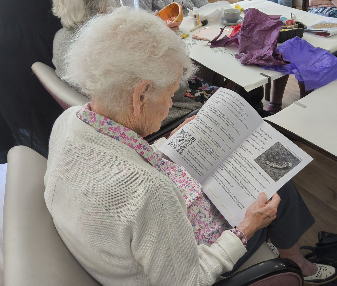 A woman in a floral blouse and light cardigan studies a printed page with image of semi-buried pendant. On the table in front of her are pieces of blue and purple tissue paper and assorted art materials