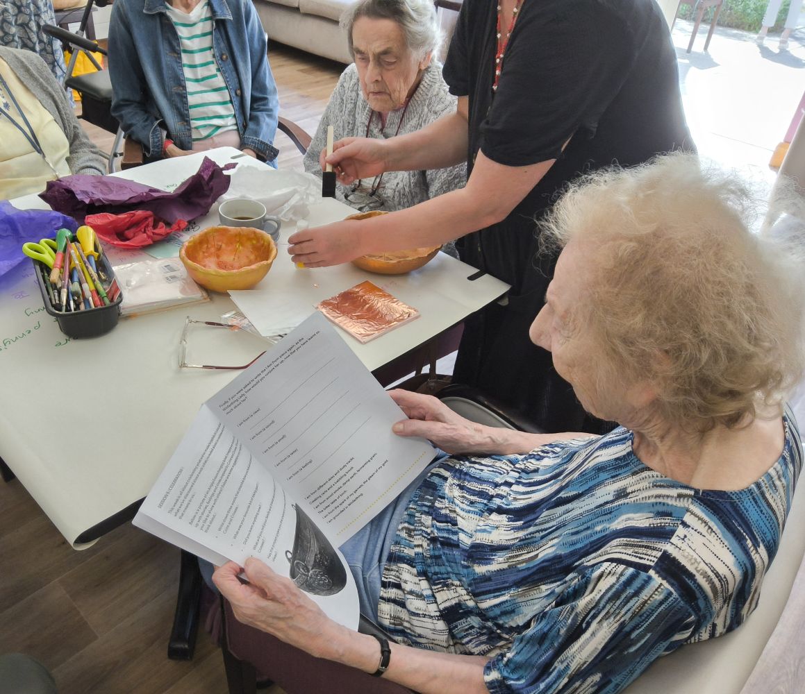 As one woman reads a publication, another looks on intently as a figure demonstrates brushing glue into a bowl before applying gold leaf
