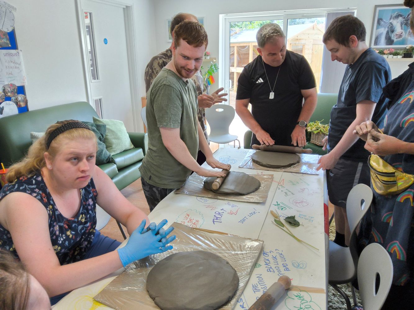 A group of people standing at a table, rolling out large pieces of clay, whilst a woman is seated in the foreground