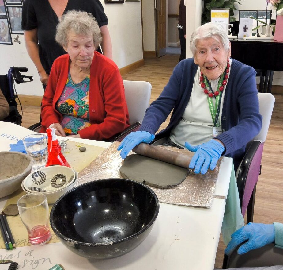 Two women seated at a table, one rolling out a slab of clay whilst the other looks on