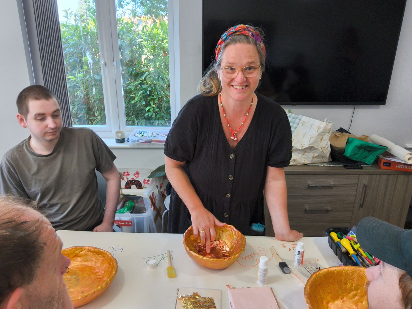A woman with a colourful head scart, necklace and dressed in black frock faces camera, her finger pushed into a gold bowl where she is applying some gold leaf. A young adult seated to her right looks on