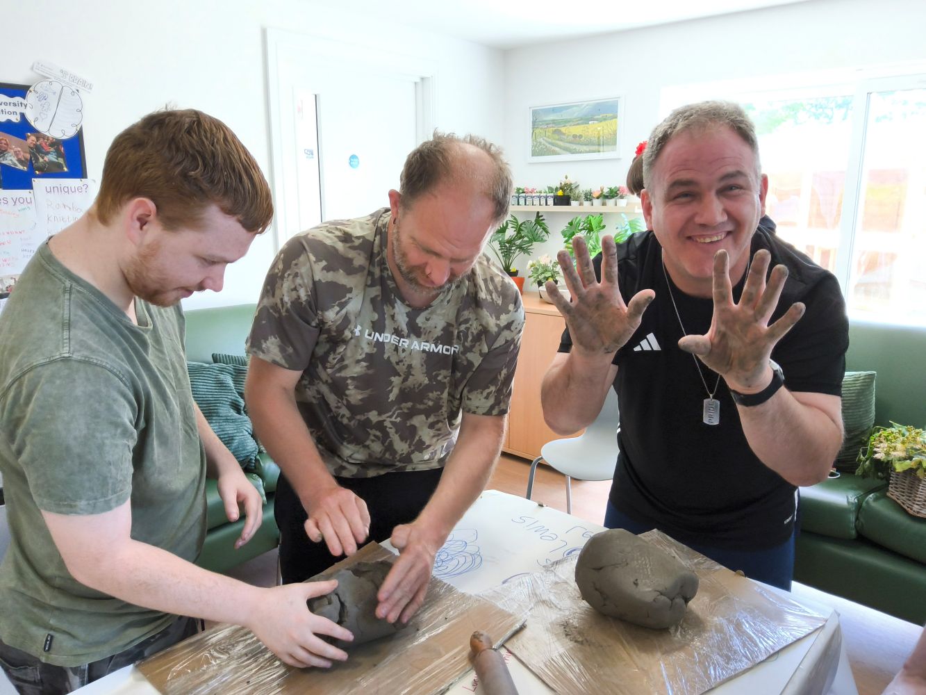 A man holds up clay-covered hands to the camera as others continue to soften up a large lump of clay