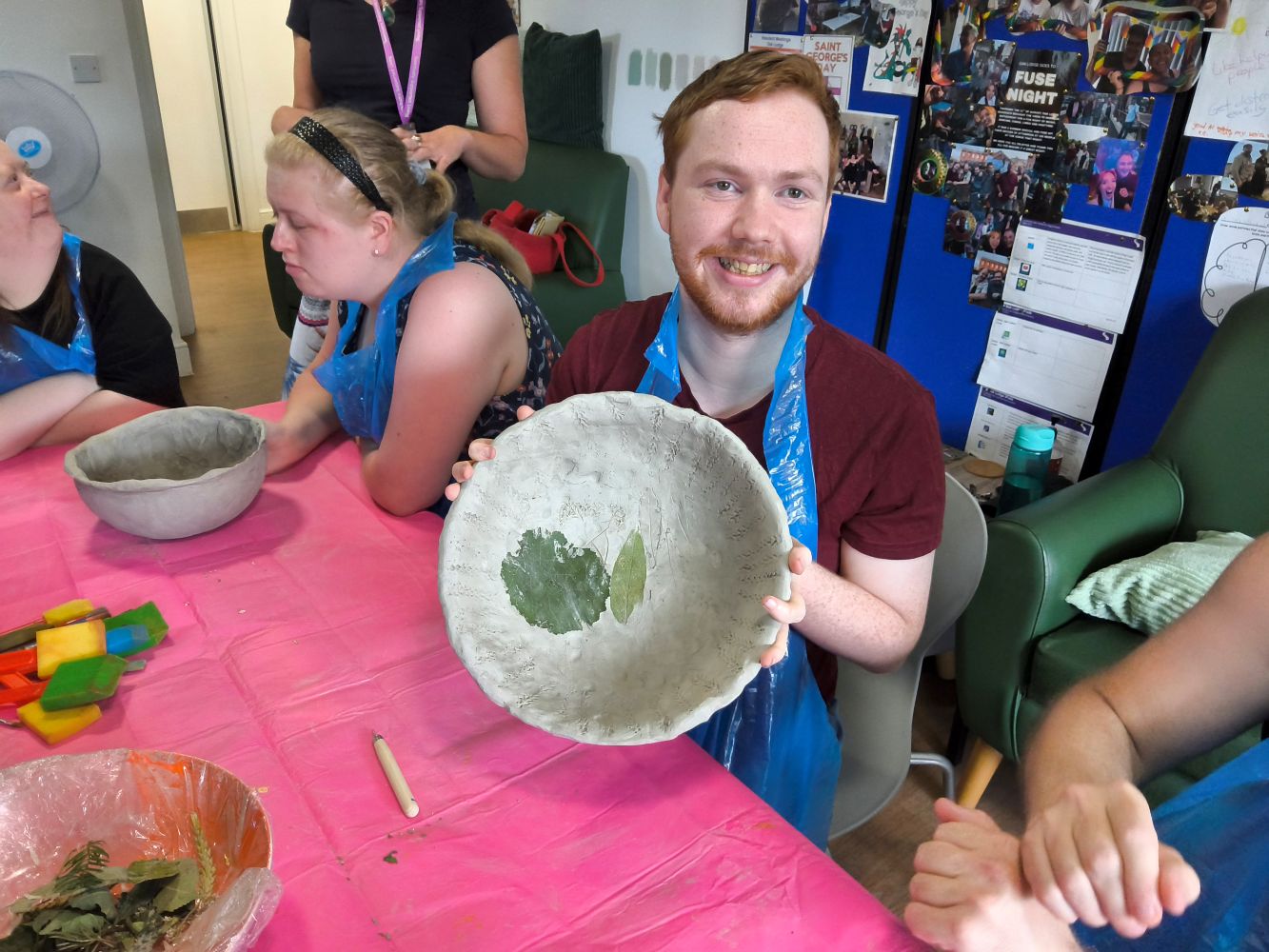 A young man holds up a clay bowl with two leaves adhered inside. There is another bowl on the table, sponges and loose foliage