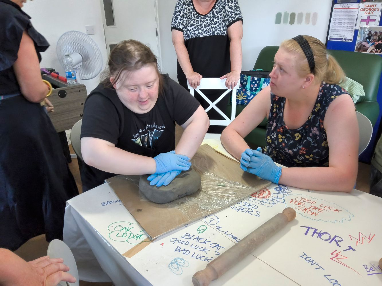 Two women seated at a table, others in the background. Women at table are wearing blud gloves and one is pushing down hard on a large lump of clay