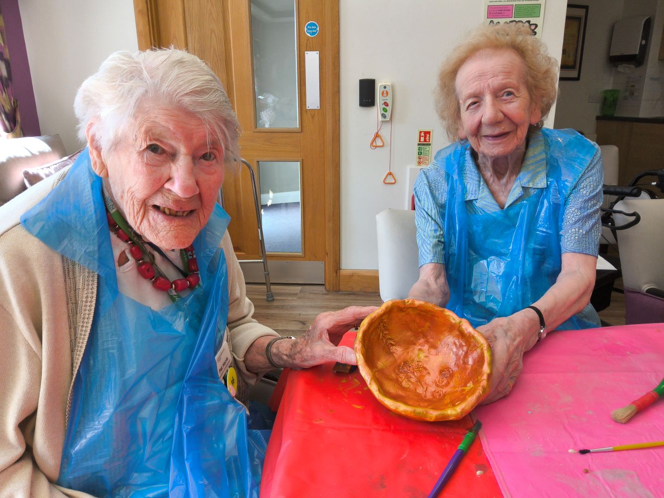 Two women wearing blue plastic aprons look to camera, holding their painted bowl to reveal the interior