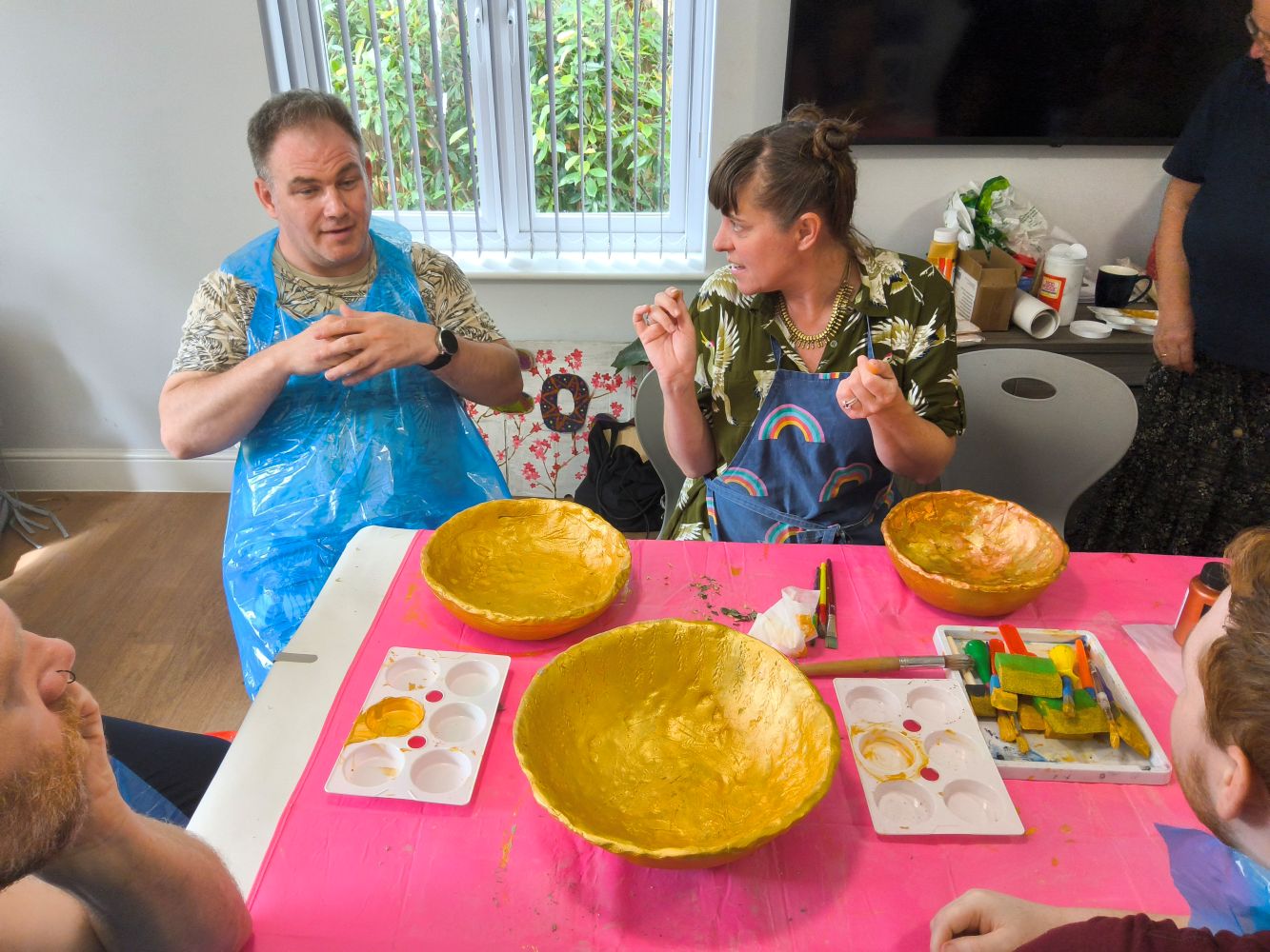 A pink covered table with 3 gold painted bowl plus palettes, paint and brushes. A pair are in discussion about something, the woman turning to face the man who is clearly considering his answer