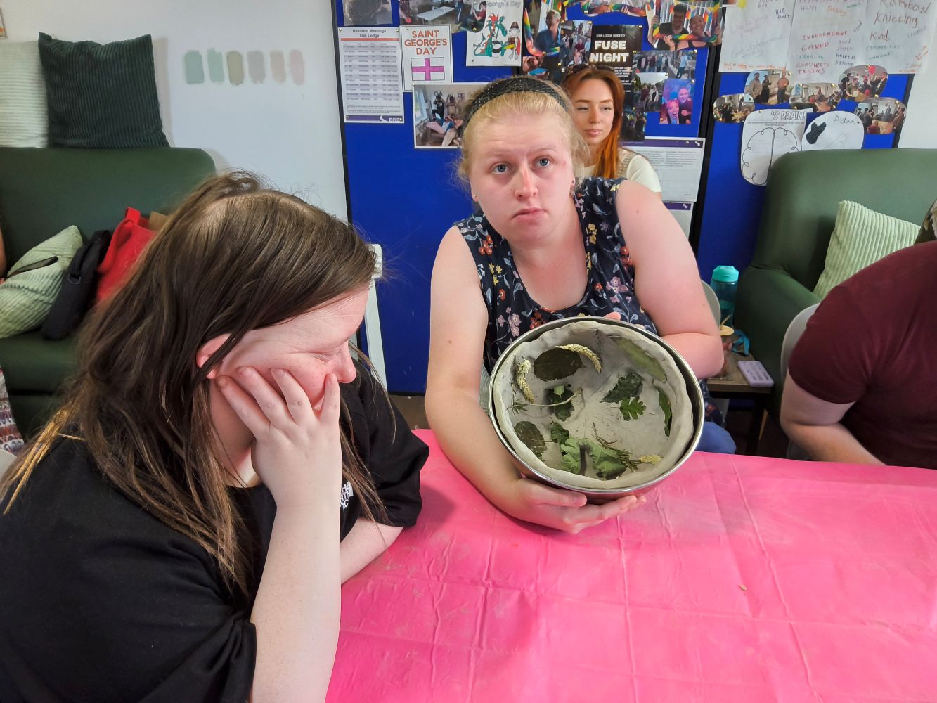 Two young women seated at a table covered in a pink cloth hold a clay bowl up for inspection. There are leaves stuck to the inside of the bowl