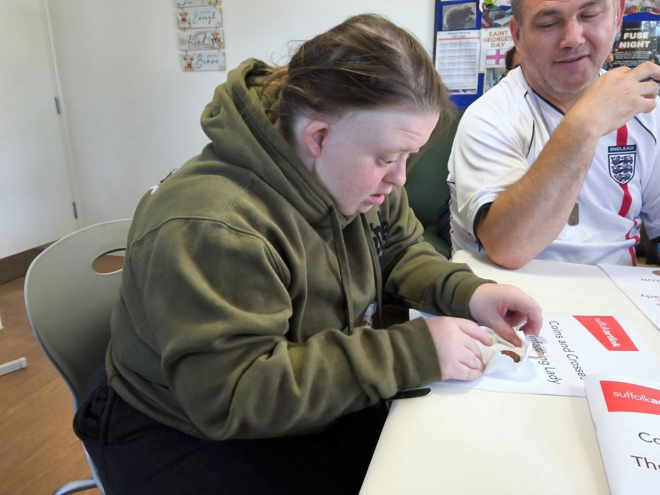 A person carefully unwrapping a small clay disc, whilst another, wearing an England football shirt, looks on.