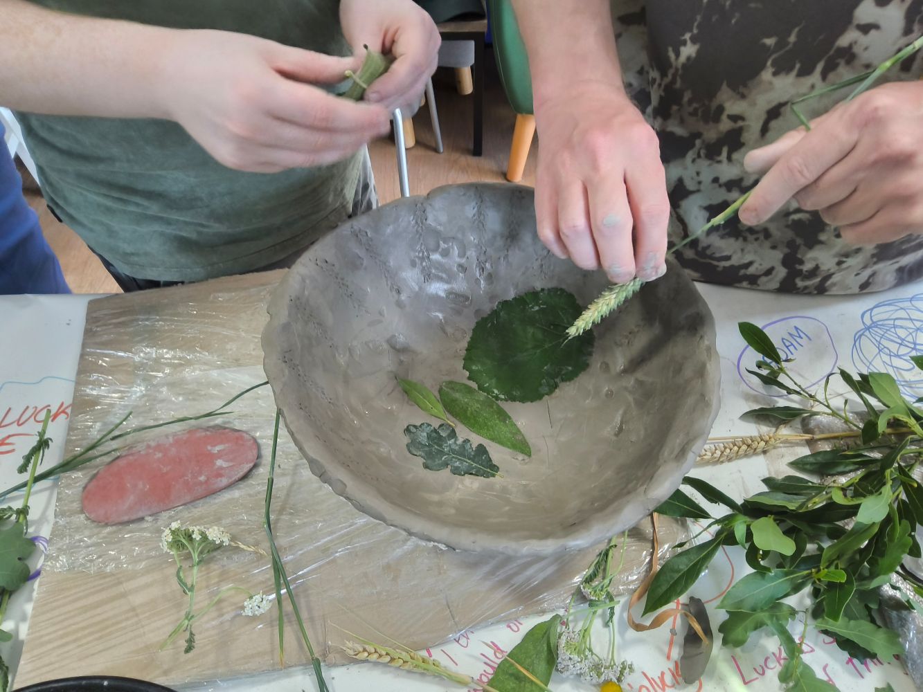 A hand holding an ear of corn over a wet clay bowl with leaves pressed inside