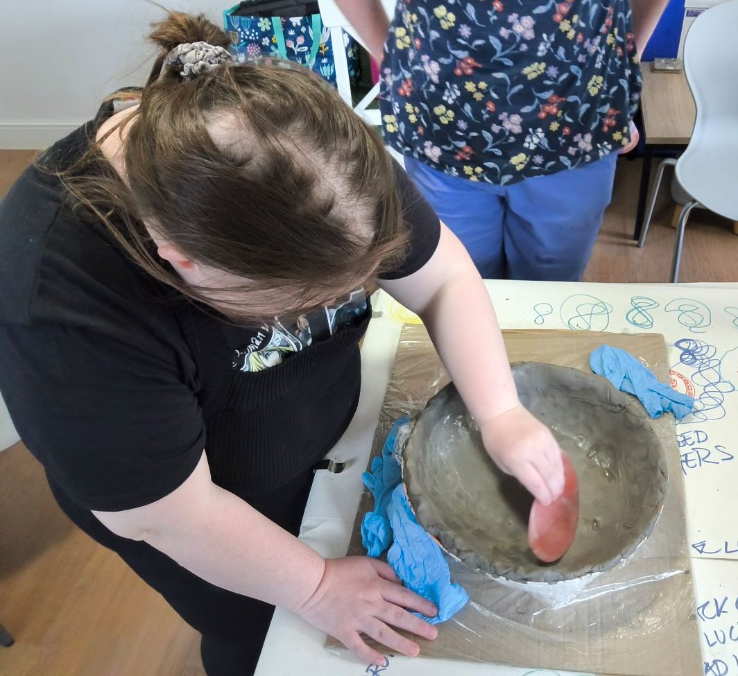 A person leans over the wet clay bowl using a rubber kidney to smooth out the interior