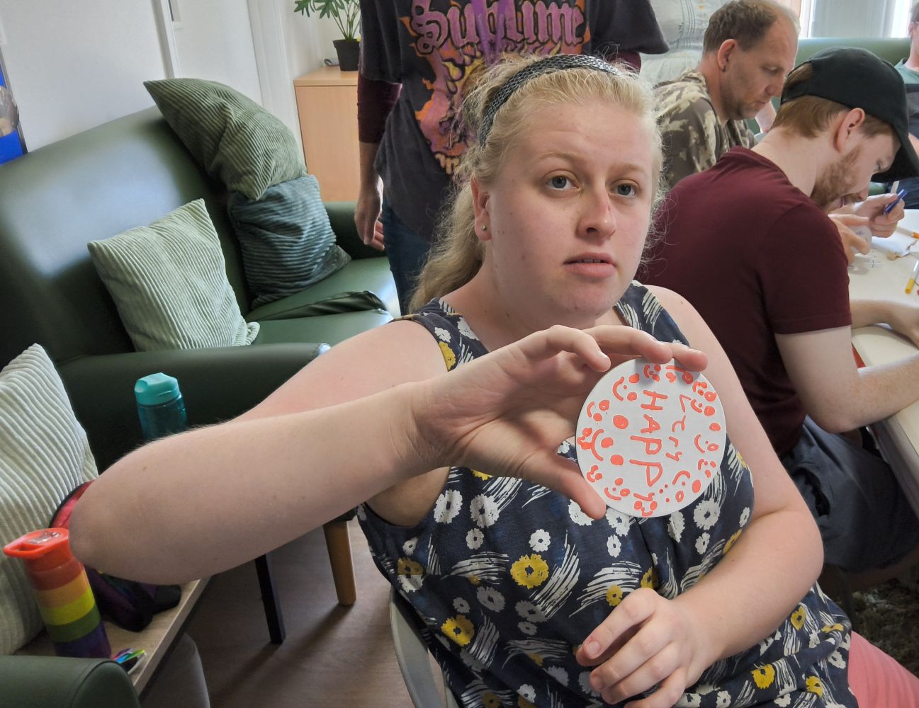 A young woman looks to camera, holding up a white card disc on which she's written Very Happy. Other people work in the background.