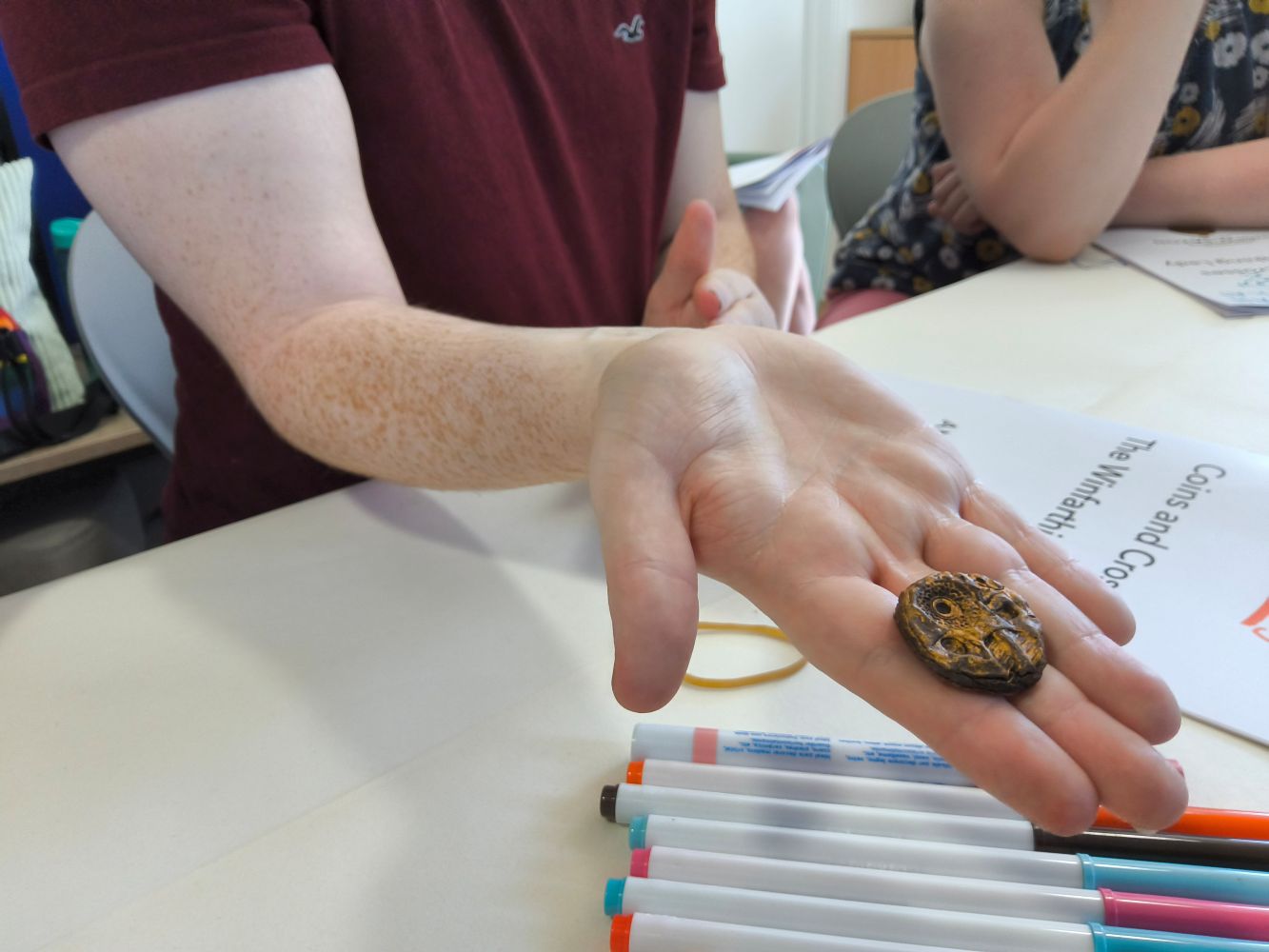 A copper disc in a person's hand, stretch towards the camera, with a row of coloured pens nearby and a publication with legible words Coins and Crosses