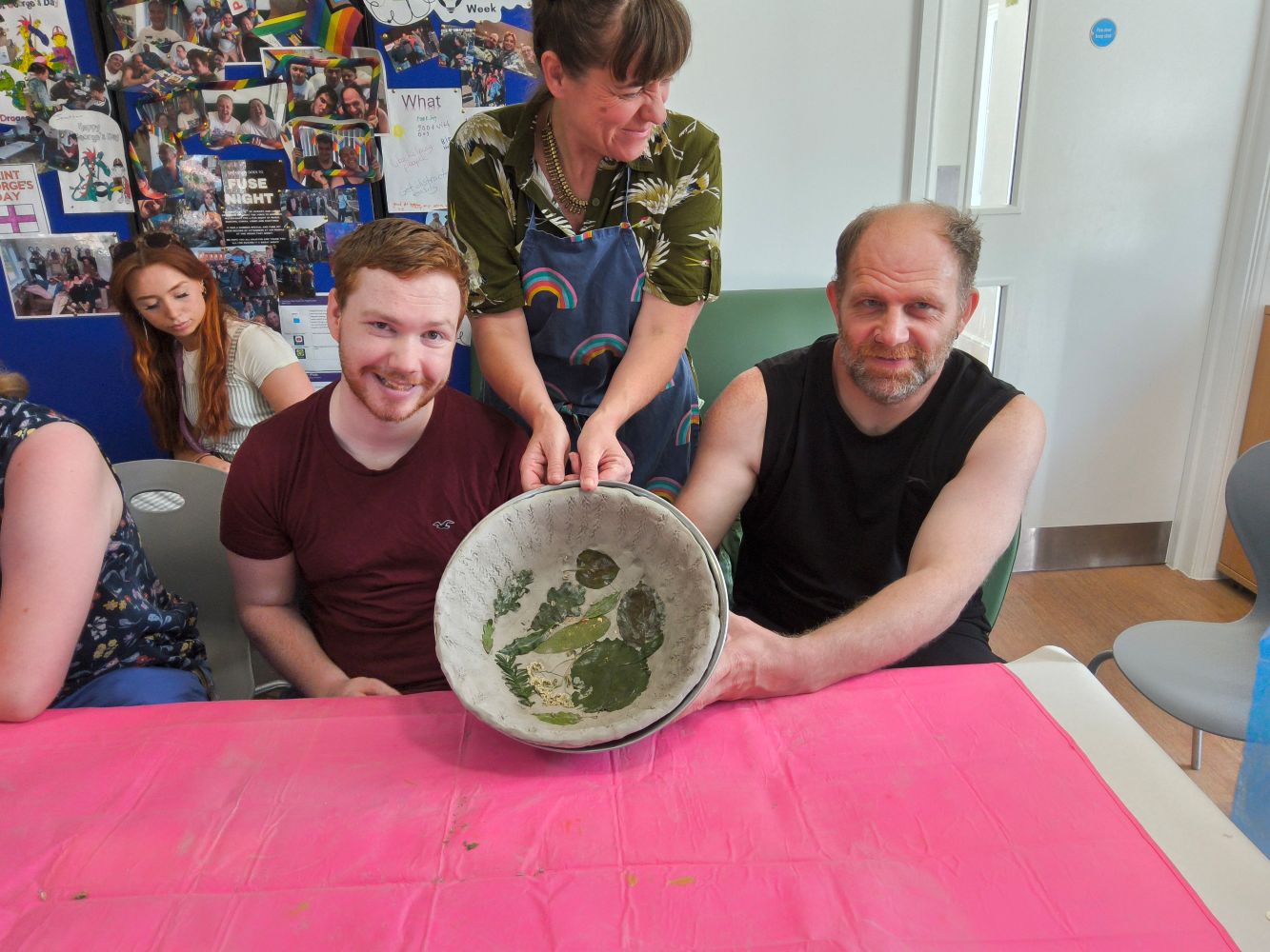 Two people seated at a pink-covered table, holding up a dried clay bowl imprinted with leaves. A woman stand behind them, helping to steady the large bowl and another woman with long auburn hair is seated in the background