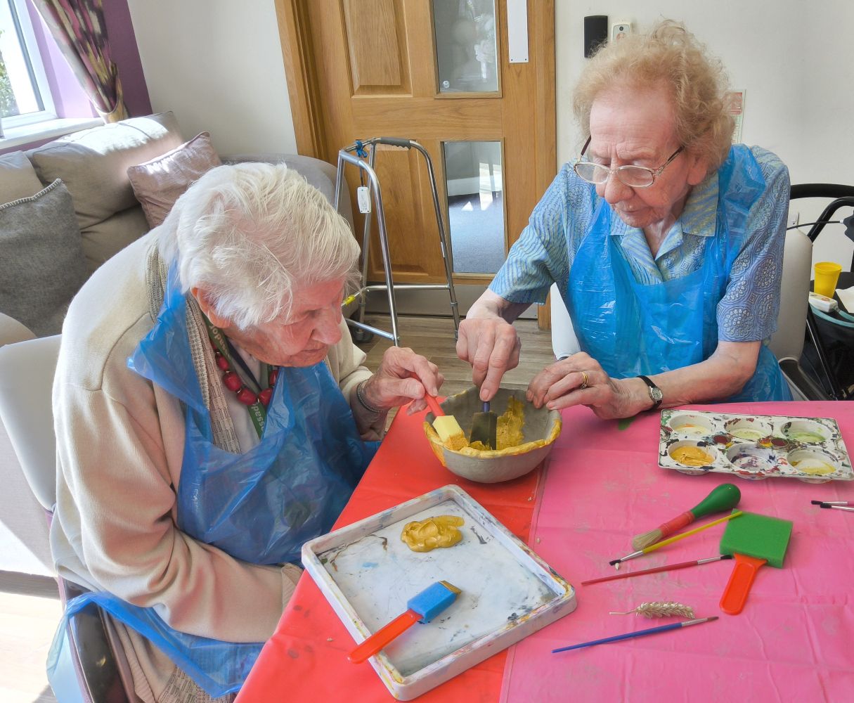 Two women wearing protective blue aprons apply gold paint to a clay bowl