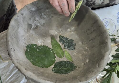 Leaves pressed into a wet clay bowl with a hand holding an ear of corn using it to decorate the rim of the bowl