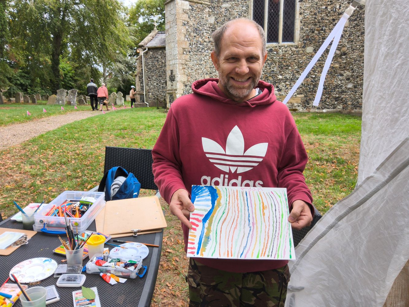 A person in a plum and white Adidas hoodie faces the camera, holding a colourful striped picture. Beside them is a table covered with paint and other art materials and in the background is a flint walled church.