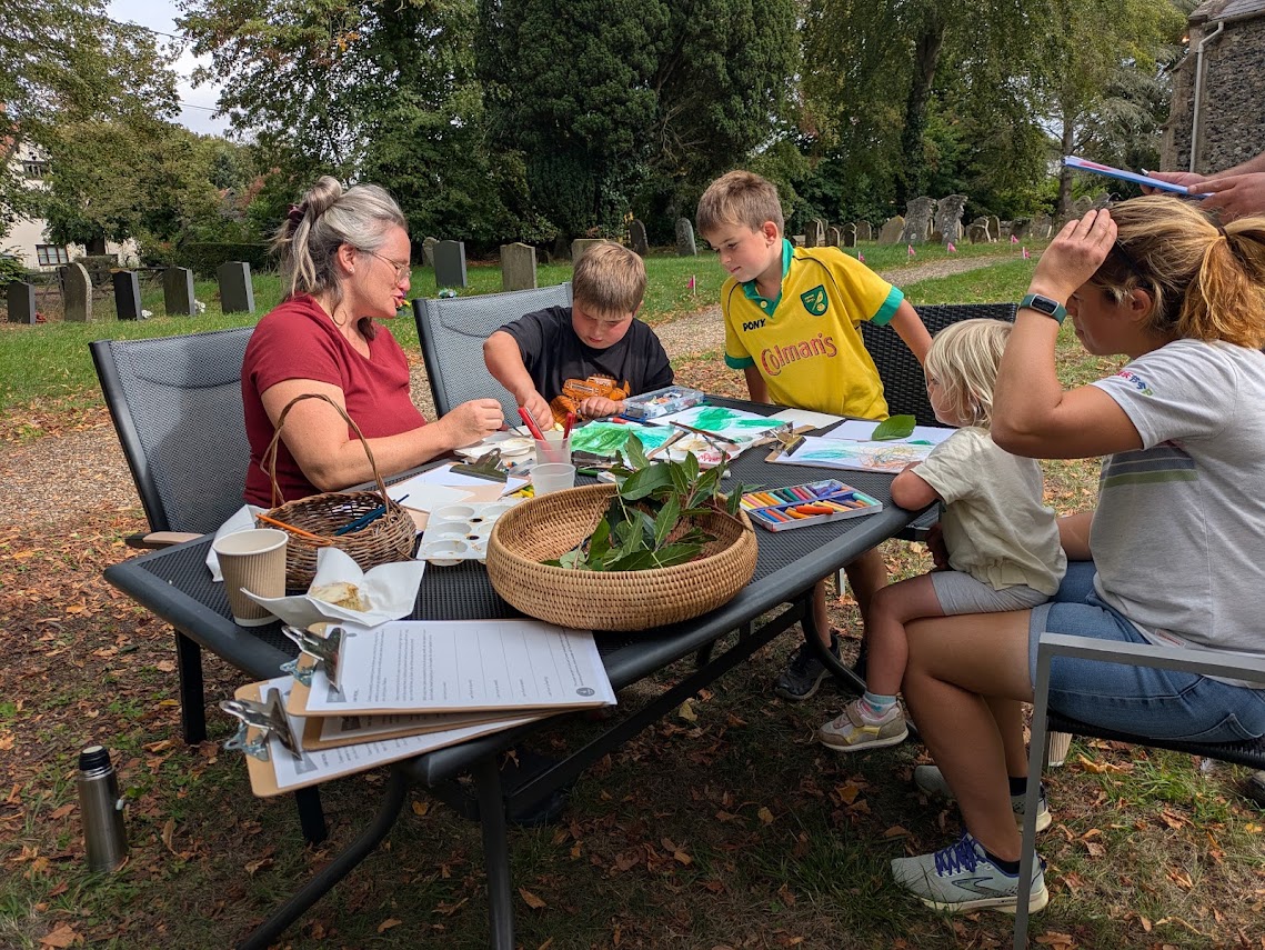 A group of children and adults gathered round a table in a churchyard. The table is covered with art materials and the children are watching intently as one of them squeezes paint out of a tube.