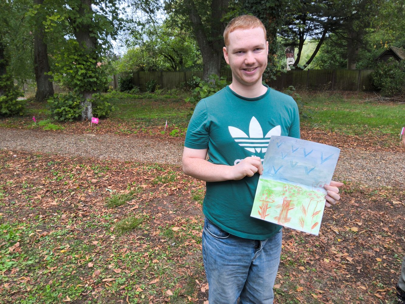 A young man grins broadly at the camera as he holds up his picture of trees and birds. In the background are several mature trees and small flags, celebrating Heritage Open Day