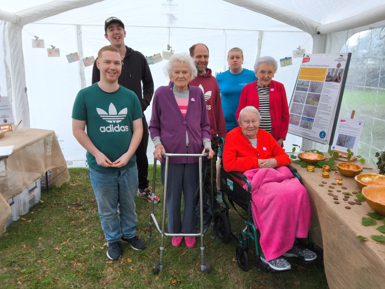 A group of adults, one of whom is seated with a pink rug covering their knees and another holds a walker, standing in a marquee with gold exhibits displayed on their left