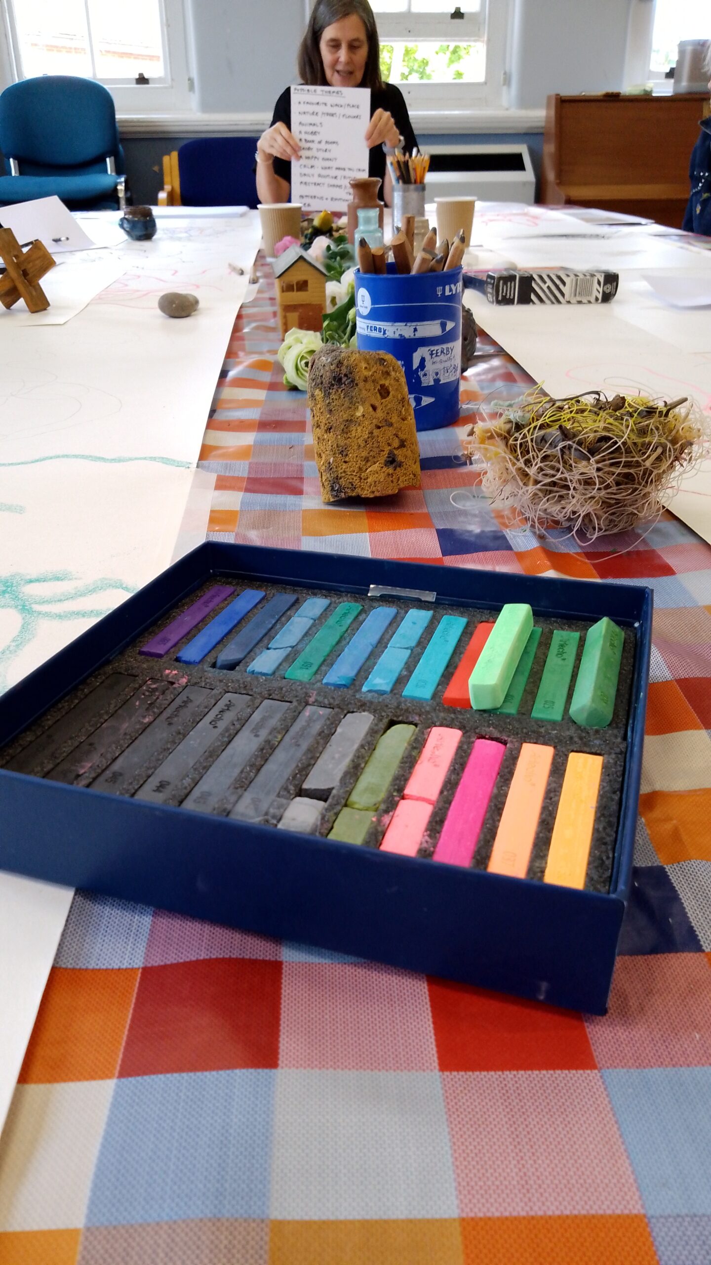 Shot looking up a table with a case of soft pastels pastels in the foreground and long pieces of paper each side an a person holding up a sheet with possible themes sitting at the head of the table.